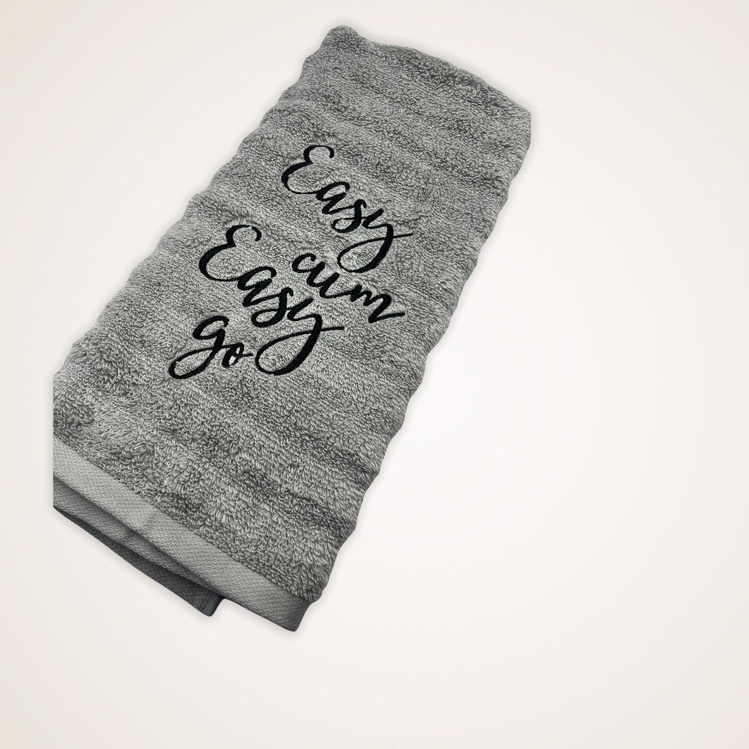 The image features an embroidered hand towel. The gray towel is adorned with Easy cum, Easy Go in black thread.  It serves as both a functional and decorative item, suitable for various settings such as bathrooms or kitchens.