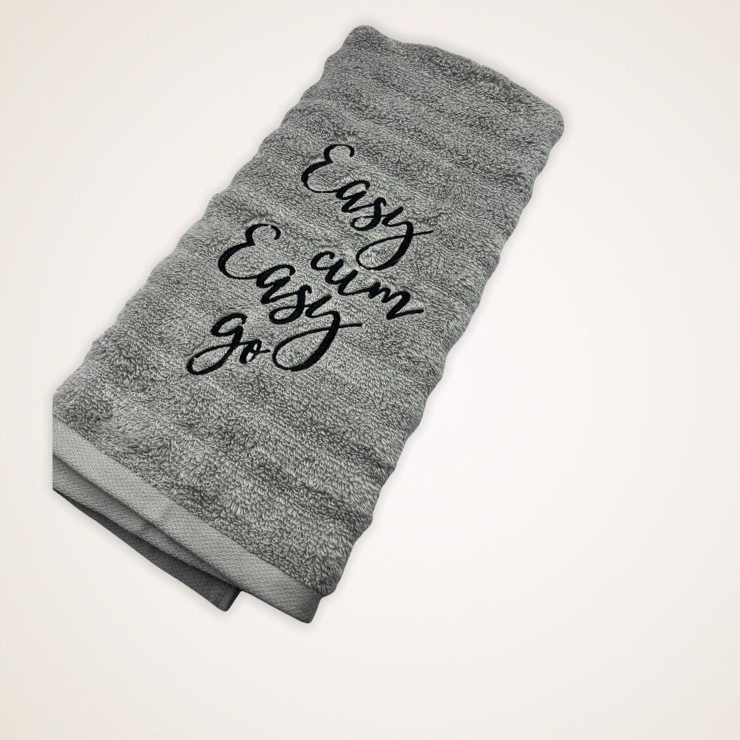 The image features an embroidered hand towel. The gray towel is adorned with Easy cum, Easy Go in black thread.  It serves as both a functional and decorative item, suitable for various settings such as bathrooms or kitchens.