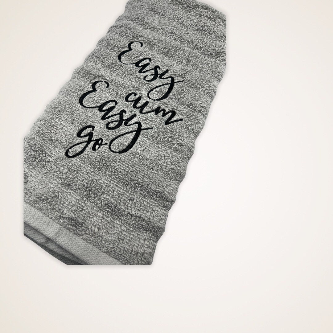 The image features an embroidered hand towel. The gray towel is adorned with Easy cum, Easy Go in black thread.  It serves as both a functional and decorative item, suitable for various settings such as bathrooms or kitchens.