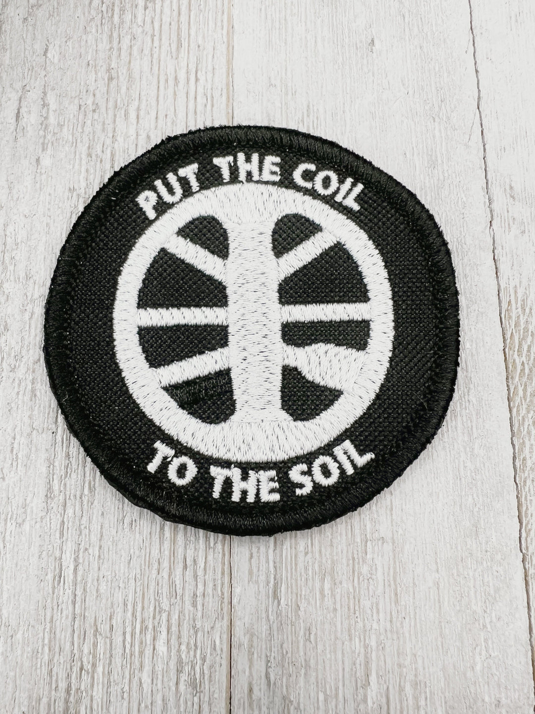 Embroidered patch featuring Put the Coil to the Soil in white on a black background. Sew on, Iron on, or hook and loop backing available. Finished with a merrow edge for a polished finish.