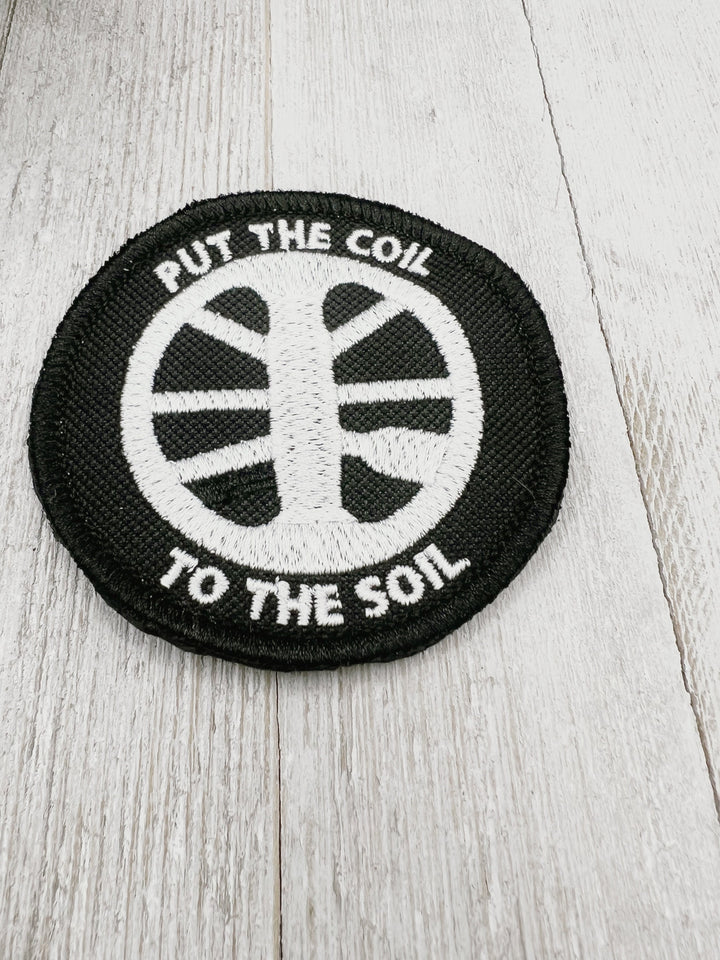 Embroidered patch featuring Put the Coil to the Soil in white on a black background. Sew on, Iron on, or hook and loop backing available. Finished with a merrow edge for a polished finish.