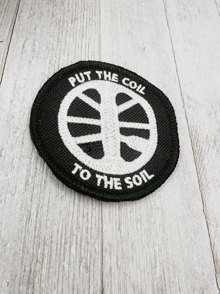 Embroidered patch featuring Put the Coil to the Soil in white on a black background. Sew on, Iron on, or hook and loop backing available. Finished with a merrow edge for a polished finish.